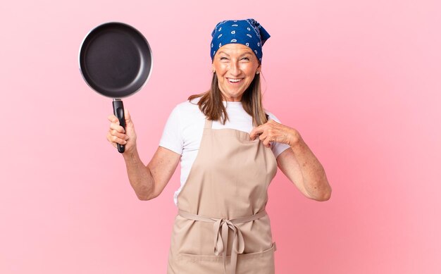 Bella donna di mezza età che si sente felice e indica se stessa con un eccitato e tiene in mano una padella. concetto di chef