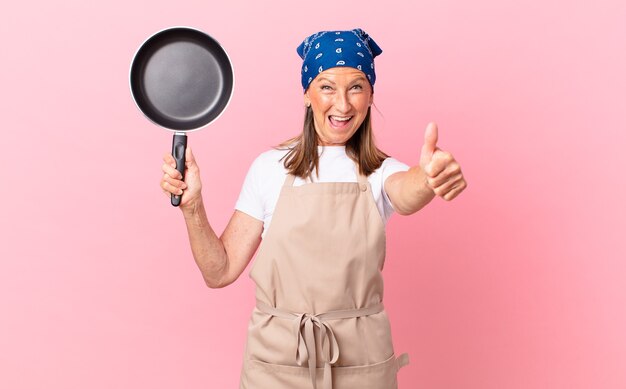 Pretty middle age woman feeling proud,smiling positively with thumbs up and holding a pan. chef concept