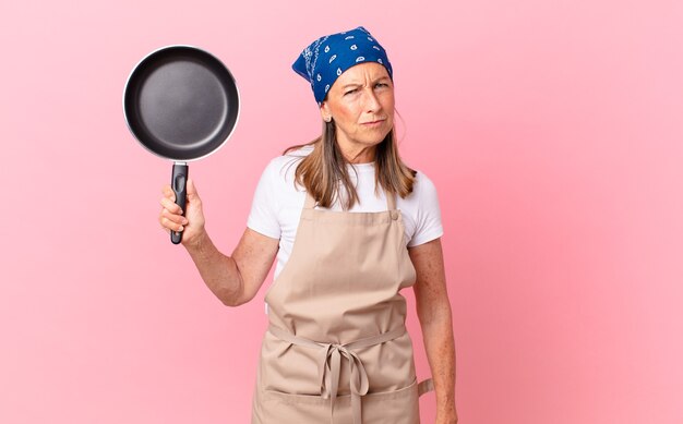 꽤 중년 여성은 슬프거나 화가 나거나 화가 나서 옆을 보고 냄비를 들고 있습니다. 요리사 개념