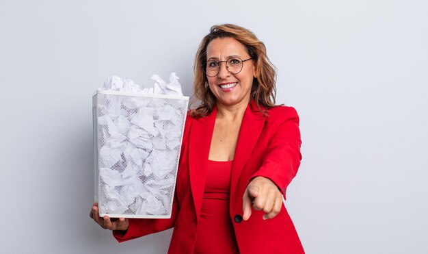 Photo pretty middle age woman pointing at camera choosing you. paper balls bin concept