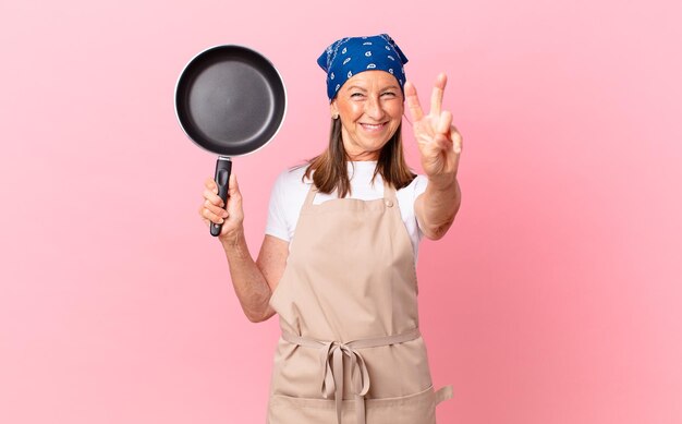 Pretty middle age woman smiling and looking friendly, showing number two and holding a pan. chef concept