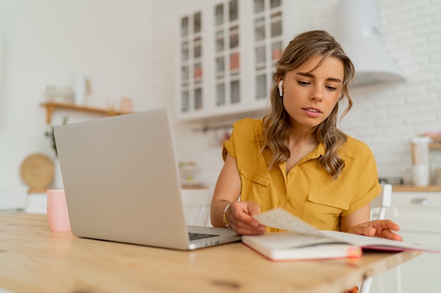 Pretty miling student woman using laptop and writing notes on her modern light kitchen.