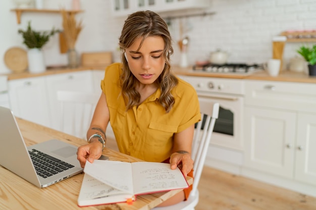 Pretty miling student woman using laptop and writing notes on her modern light kitchen