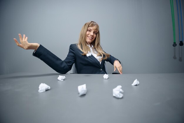 Pretty millennial blonde businesswoman worker in blue jacket\
throws papers into the camera sitting at workplace in gray modern\
office big boss concept copy space wide angle