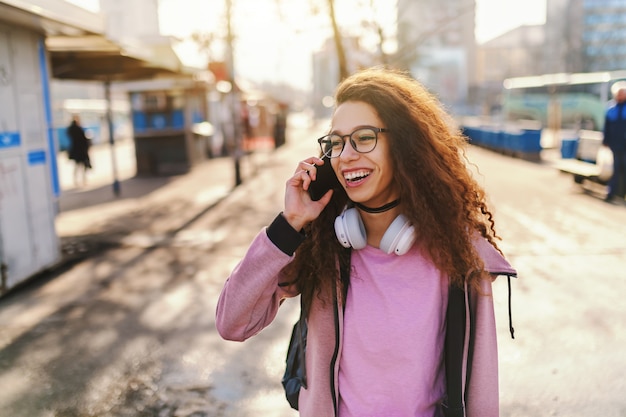 Pretty mixed race teenage hipster girl with toothy smile and backpack on backs talking on the phone while standing on the street.
