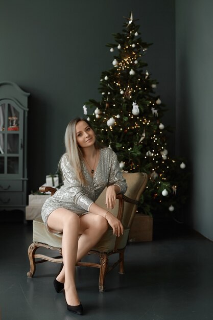 A pretty model girl with platinum blond hair wearing a modish dress sits next to a christmas tree in...