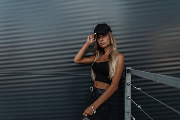 Pretty model woman with black swag clothes summer top and jeans shorts in fashion black mockup cap  stands near a dark metal wall