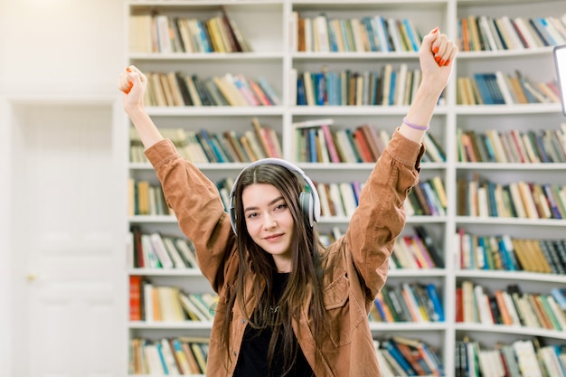 Pretty modern confident 25-aged girl with long brown hair listening favourite music in earphones, holding arms up and posing near the bookshelves in the library