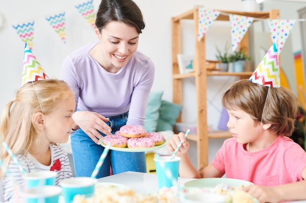 Красивая мама одного из маленьких детей держит пончики над сервированным столом, показывая их друзьям на вечеринке по случаю дня рождения