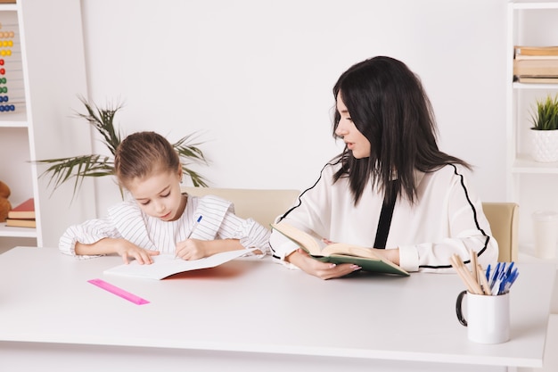 Pretty mother with cute daughter sitting with book in the room at home.