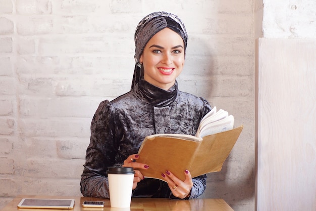 Pretty Muslim woman holds a book and poses for camera in cafe