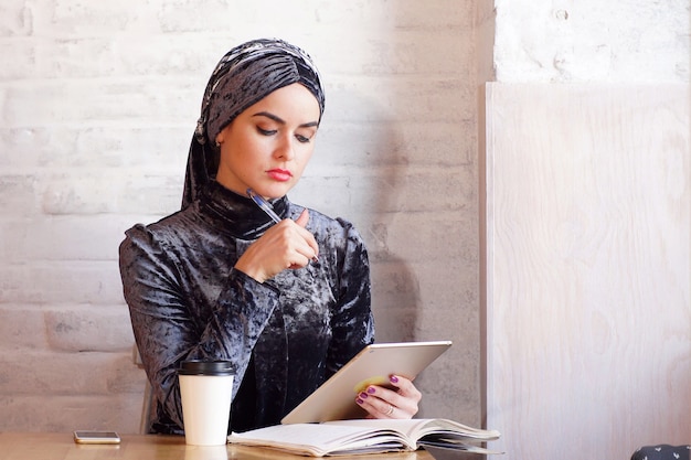 Photo pretty muslim woman holds a tablet in her hands while sitting in cafe