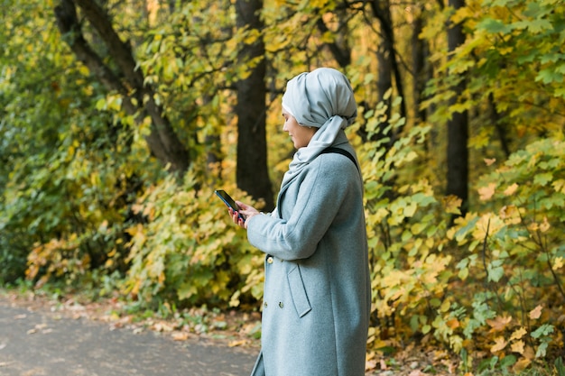 屋外で携帯電話を使用しているかなりイスラム教徒の女性。スマートフォンを使用してヒジャーブを身に着けているアラビア語の女性。都市公園でメッセージ電話にテキストメッセージを送るイスラムの少女。コピースペース