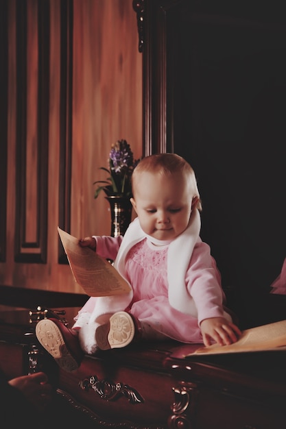 Pretty one-year-old girl in pink dress with an old book is sitting on sideboard by mirror. Child in library interior. Concept of education and child rearing. Background for site or banner. Copy space