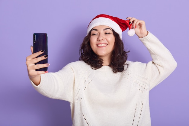 Signora piuttosto felicissima che fa selfie, indossa un maglione lavorato a maglia e cappello da babbo natale, posa isolato sul muro lilla, femmina che tiene smart phone e guarda lo schermo del dispositivo con un sorriso felice.