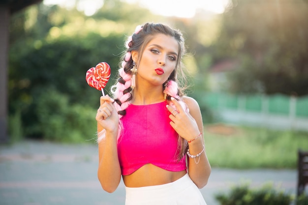 Красивая и позитивная девушка в розовом топе держит конфетное сердце на палочке.