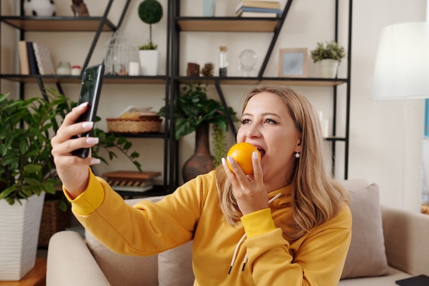 Pretty positive young blond woman biting unpeeled orange and taking selfie for social media