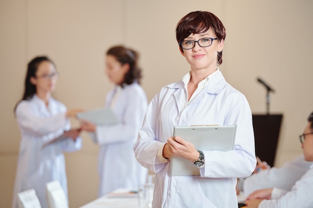 Pretty positive young doctor in labcoat standing with clipboard in hands, her collagues talking in background