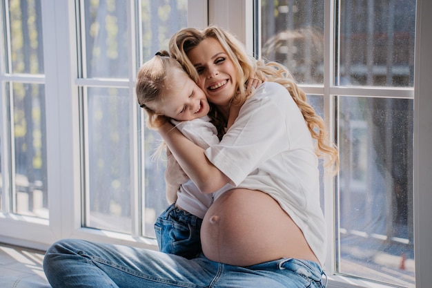 Pretty pregnant caucasian woman with daughter in white tshirt and blue jeans sits on floor