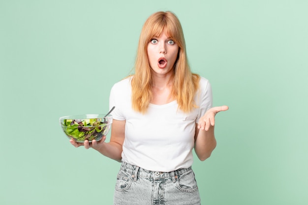 Pretty red head woman amazed, shocked and astonished with an unbelievable surprise and holding a salad. diet concept