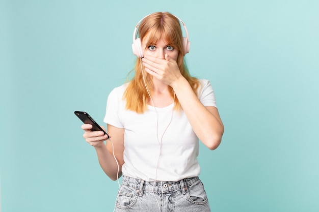 Pretty red head woman covering mouth with hands with a shocked and listening music with headphones