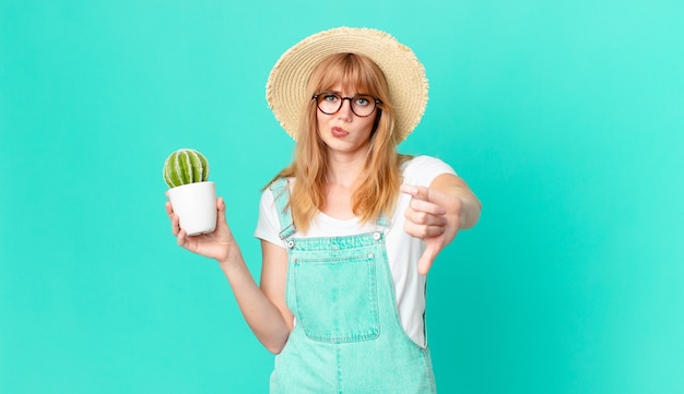 Pretty red head woman feeling cross,showing thumbs down and holding a potted cactus. farmer concept