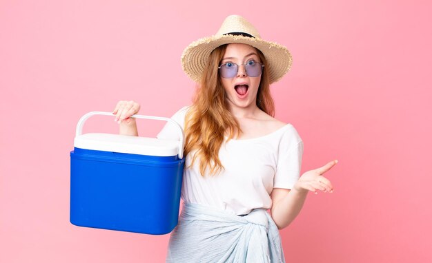 Pretty red head woman feeling extremely shocked and surprised and holding a picnic portable refrigerator