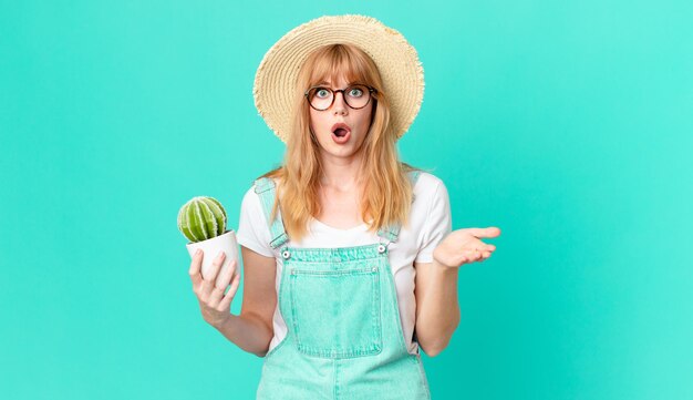 Pretty red head woman feeling extremely shocked and surprised and holding a potted cactus. farmer concept