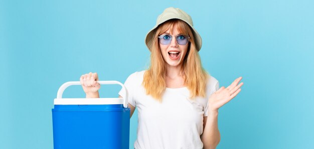 Pretty red head woman feeling happy and astonished at something unbelievable and holding a portable refrigerator. summer concept