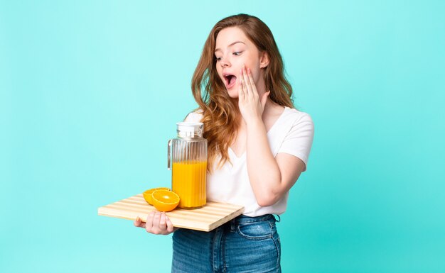 Pretty red head woman feeling happy, excited and surprised and holding an orange juice