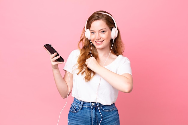 Pretty red head woman feeling happy and facing a challenge or
celebrating with headphones and smartphone