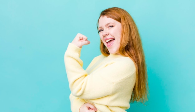 Pretty red head woman feeling happy satisfied and powerful flexing fit and muscular biceps looking strong after the gym