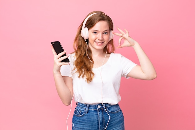 Pretty red head woman feeling happy, showing approval with okay gesture with headphones and smartphone
