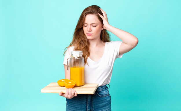 Pretty red head woman feeling puzzled and confused, scratching head and holding an orange juice