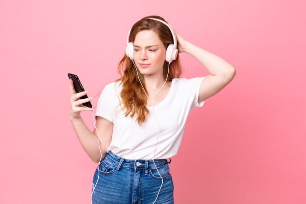 Pretty red head woman feeling puzzled and confused, scratching head with headphones and smartphone