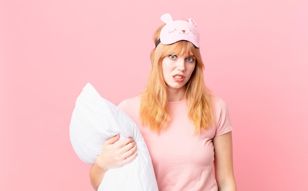 Pretty red head woman feeling puzzled and confused. wearing pajamas and holding a pillow