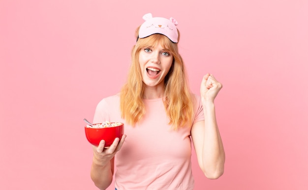 Pretty red head woman feeling shocked,laughing and celebrating success wearing pajamas and holding a flakes bowl