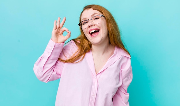 Pretty red head woman feeling successful and satisfied smiling with mouth wide open making okay sign with hand