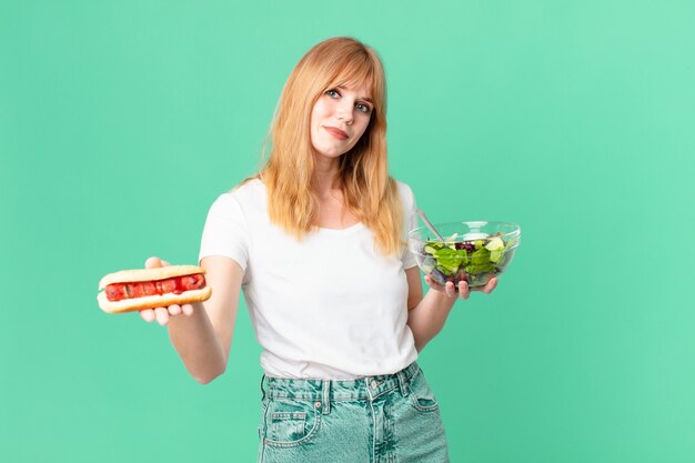 Pretty red head woman holding a salad and a hot dog. diet concept