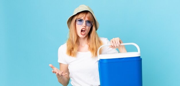 Foto bella donna dalla testa rossa che sembra arrabbiata, infastidita e frustrata e che tiene in mano un frigorifero portatile. concetto di estate