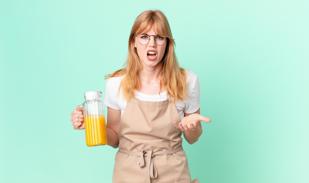 Pretty red head woman looking angry annoyed and frustrated with apron preparing an orange juice
