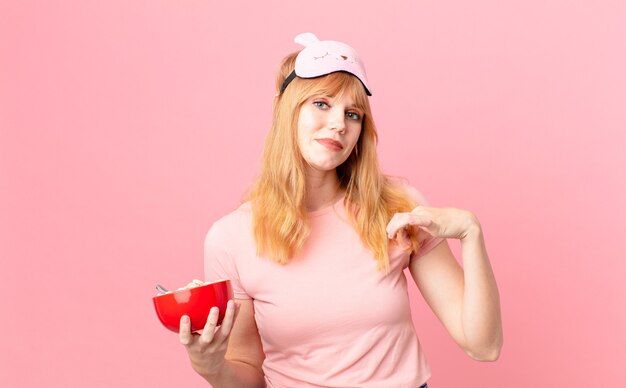 Pretty red head woman looking arrogant, successful, positive and proud wearing pajamas and holding a flakes bowl