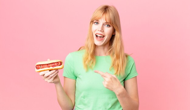 Pretty red head woman looking excited and surprised pointing to the side and holding a hot dog