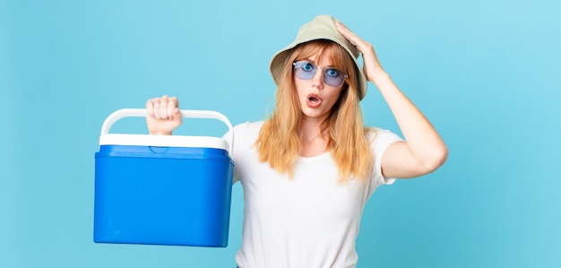 Pretty red head woman looking happy, astonished and surprised and holding a portable refrigerator. summer concept