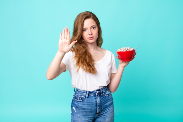Pretty red head woman looking serious showing open palm making stop gesture and holding a breakfast bowl