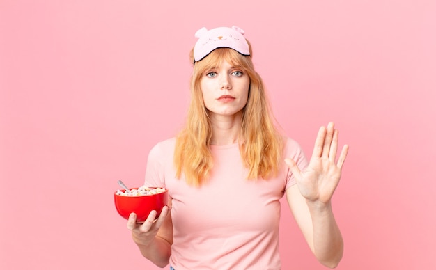 Pretty red head woman looking serious showing open palm making stop gesture wearing pajamas and holding a flakes bowl