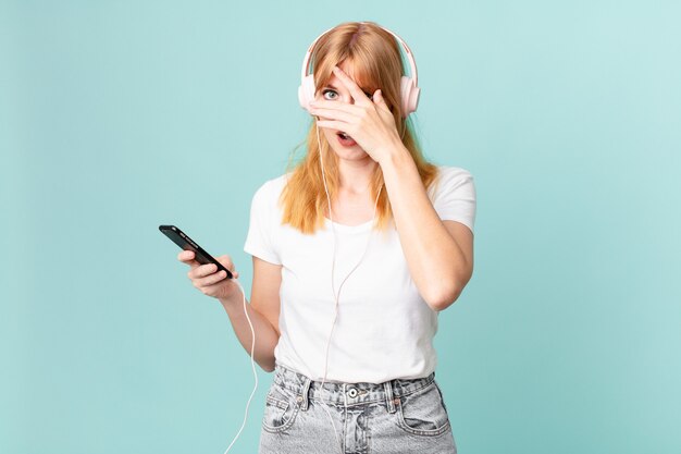 Pretty red head woman looking shocked, scared or terrified, covering face with hand and listening music with headphones