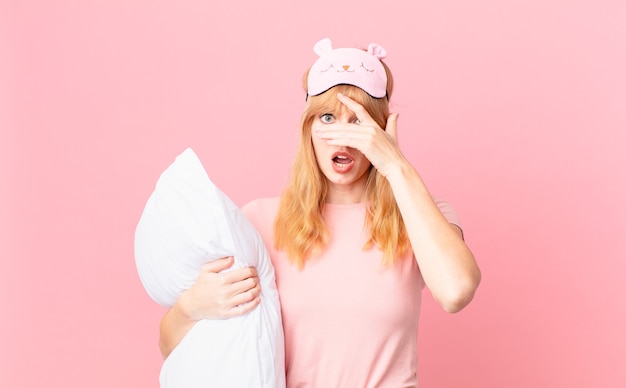 Pretty red head woman looking shocked, scared or terrified, covering face with hand. wearing pajamas and holding a pillow