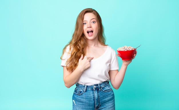 Pretty red head woman looking shocked and surprised with mouth wide open, pointing to self and holding a breakfast bowl