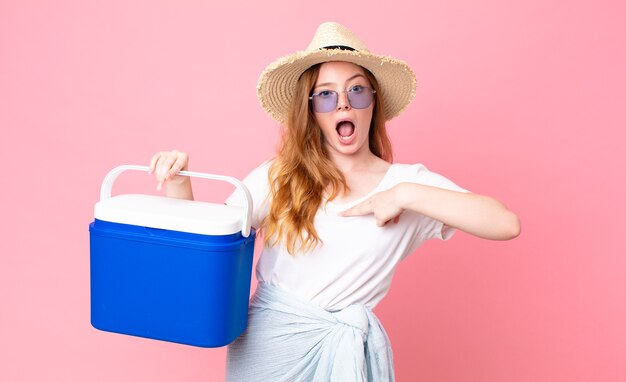 Pretty red head woman looking shocked and surprised with mouth wide open, pointing to self and holding a picnic portable refrigerator